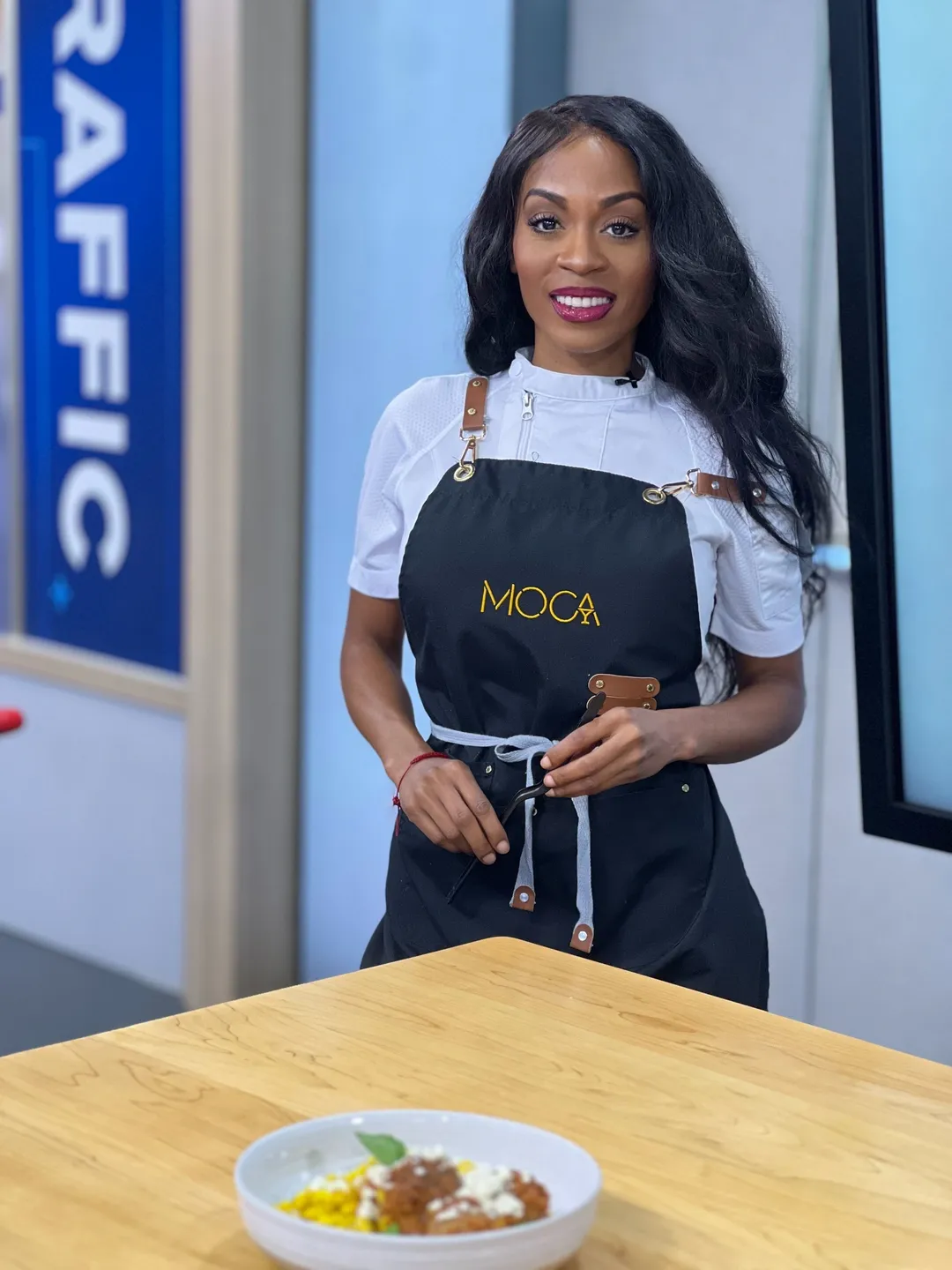 Smiling chef in MOCA apron, food on table.